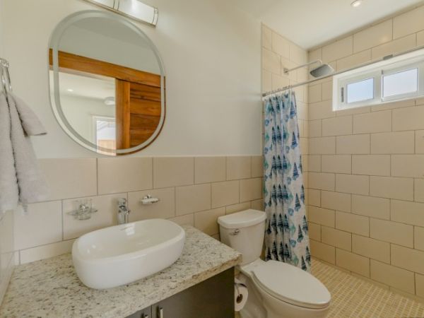 The image shows a bathroom with a round mirror, white sink on a granite countertop, a toilet, and a shower area with a blue patterned curtain.