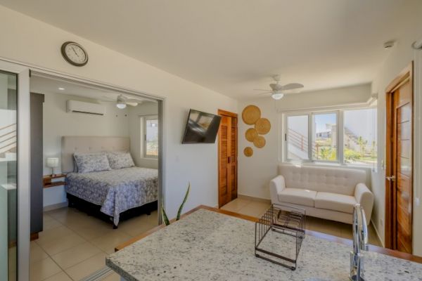 A modern interior featuring a bedroom, a living room with a white sofa, a wall-mounted TV, ceiling fans, and a granite kitchen counter with a sink.