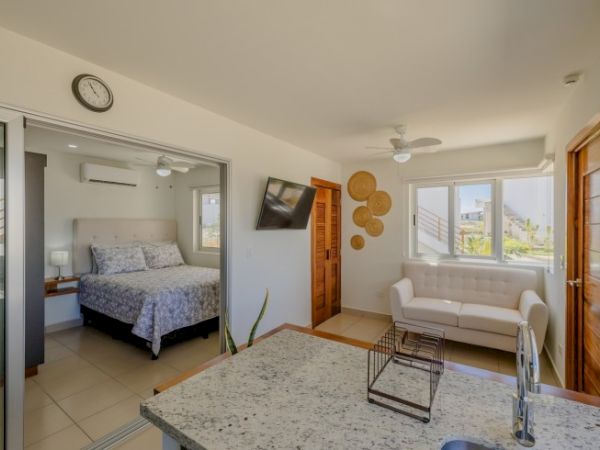 A modern interior featuring a bedroom, a living room with a white sofa, a wall-mounted TV, ceiling fans, and a granite kitchen counter with a sink.