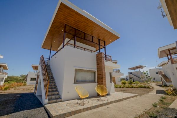 A modern, two-story white house with an external staircase, large windows, and a wooden canopy roof. Yellow chairs are placed outside.