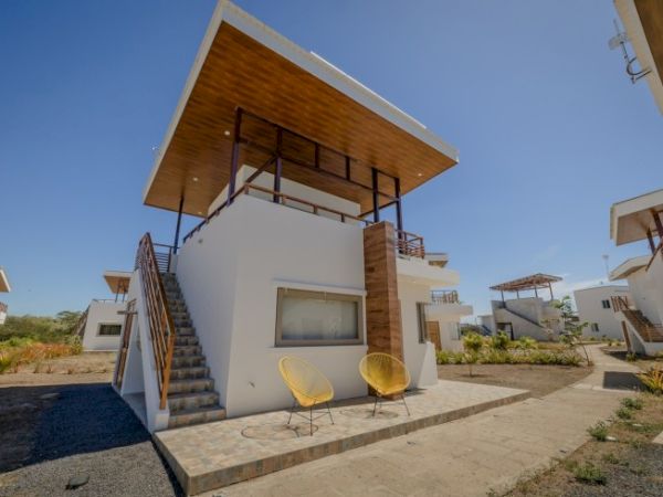 A modern, two-story white house with an external staircase, large windows, and a wooden canopy roof. Yellow chairs are placed outside.