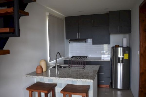 The image shows a modern kitchen with dark cabinets, a countertop with two stools, a stainless steel refrigerator, and stairs on the left.