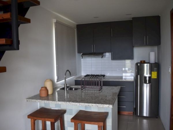 The image shows a modern kitchen with dark cabinets, a countertop with two stools, a stainless steel refrigerator, and stairs on the left.