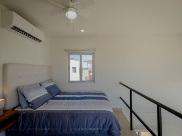 The image shows a modern bedroom with a bed, blue bedding, a headboard, a window, an air conditioner, and a ceiling fan, adjacent to a staircase.