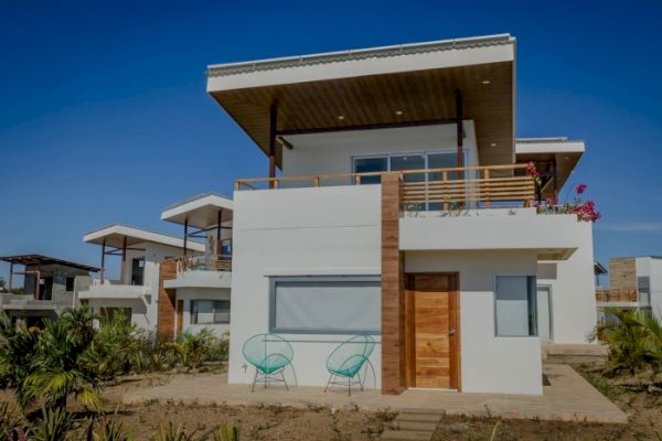 A modern, two-story house with white walls, wooden accents, and a flat roof, surrounded by greenery and featuring outdoor seating.