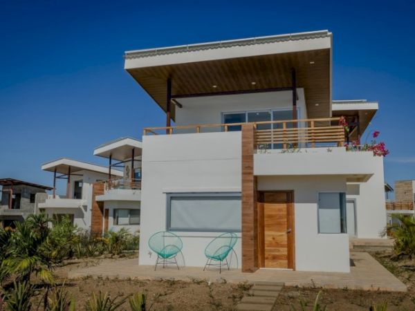 A modern, two-story house with white walls, wooden accents, and a flat roof, surrounded by greenery and featuring outdoor seating.