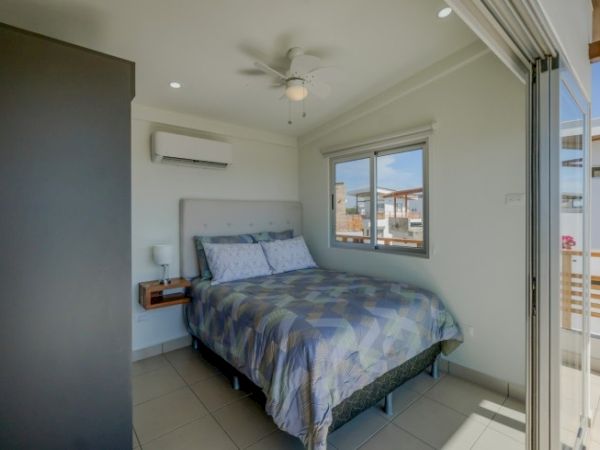 The image shows a modern bedroom with a queen-size bed, a window, air conditioning, a ceiling fan, and a sliding door leading to a balcony area.