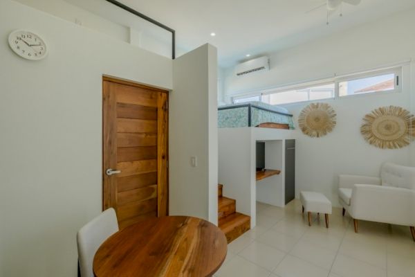 A modern, cozy room with a lofted bed, white furniture, wooden door, small round dining table, decorative wall art, a clock, and an air conditioner.