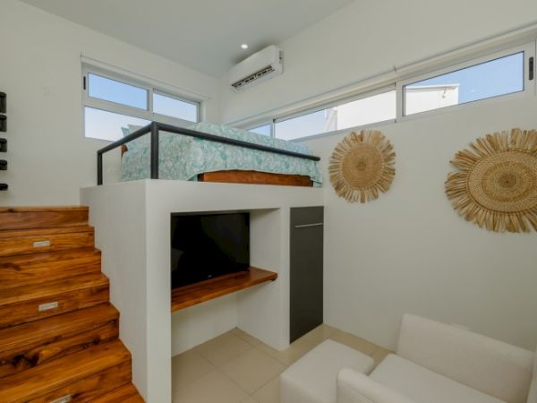 A modern loft bedroom with wooden stairs, a bed on a platform, a small seating area, wall decorations, and large windows.