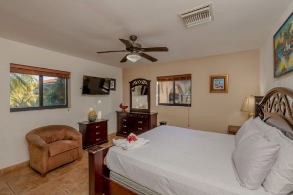 A cozy bedroom featuring a double bed, wooden furniture, a wall-mounted TV, armchair, ceiling fan, windows, and decorative wall pictures ends the sentence.