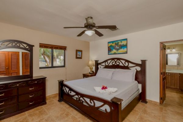 A bedroom with a large wooden bed, dresser, ceiling fan, and a bedside table. There is a window, art on the wall, and an open door to a bathroom.