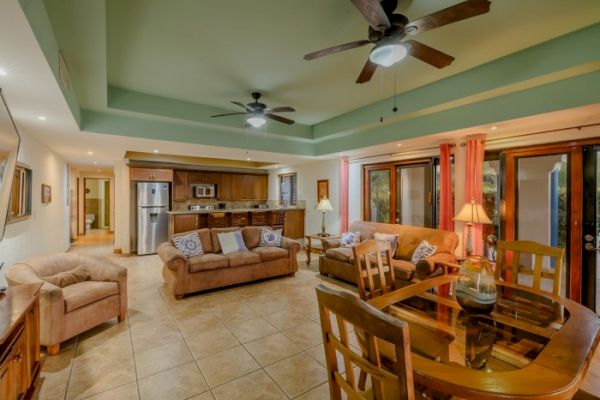 A cozy living room with beige sofas, ceiling fans, a dining table, open kitchen, and glass doors with curtains leading outside.