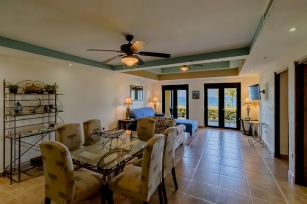 A spacious living room with a dining area, ceiling fan, and ocean view through large windows. Decor includes plants, a blue couch, and a flat-screen TV.