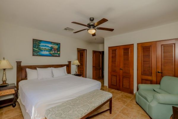 A cozy bedroom with a large bed, ceiling fan, artwork, green chair, two side tables with lamps, and wooden closet doors.