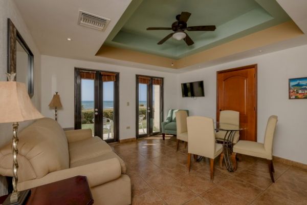 This image shows a living room with a cream-colored sofa, dining table and chairs, ceiling fan, wall-mounted TV, and a view of the ocean through windows.