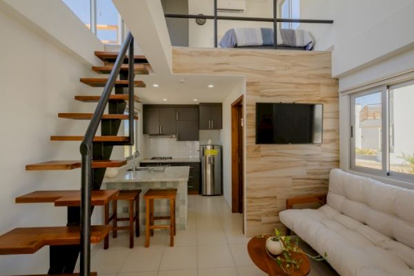 A modern loft-style apartment with a wooden staircase, small kitchen, sofa, wall-mounted TV, and upstairs bed area, elegantly decorated and well-lit.