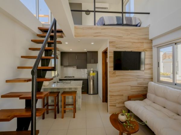 A modern loft-style apartment with a wooden staircase, small kitchen, sofa, wall-mounted TV, and upstairs bed area, elegantly decorated and well-lit.