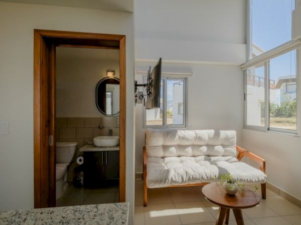The image shows a living area with a white futon, a small round table, large windows, and a bathroom with a sink and toilet to the left.