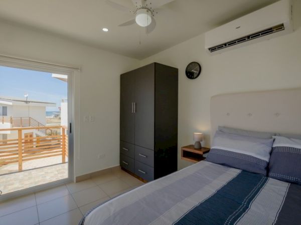 A modern bedroom with a bed, wardrobe, nightstand, ceiling fan, and air conditioner, leading to a balcony through a sliding glass door.