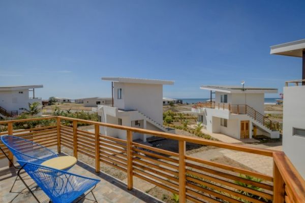 The image shows a resort-like setting with modern white buildings, balconies, and outdoor seating, with an ocean visible in the distance.