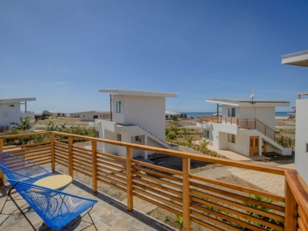The image shows a resort-like setting with modern white buildings, balconies, and outdoor seating, with an ocean visible in the distance.