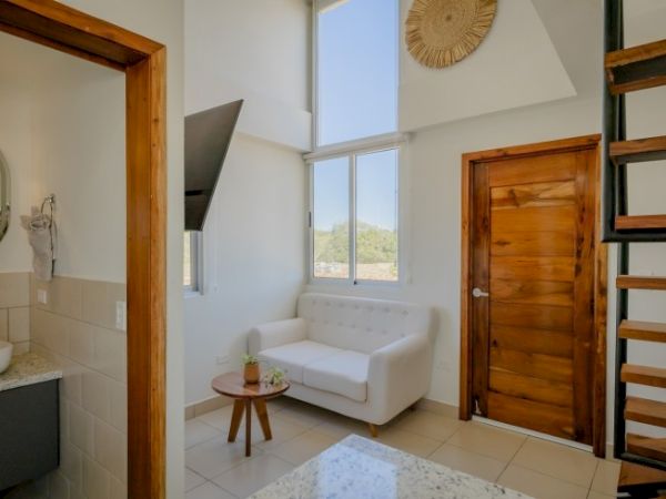 The image shows a brightly lit room with a white sofa, a wooden door, stairs, a small table, and part of a bathroom with a sink and mirror.