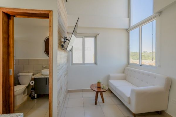 A small, bright living area with a white couch, coffee table, and wall-mounted TV, adjacent to a bathroom with a toilet and sink.