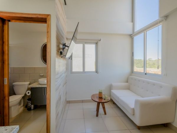 A small, bright living area with a white couch, coffee table, and wall-mounted TV, adjacent to a bathroom with a toilet and sink.