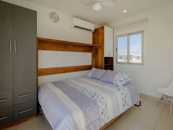 A small, modern bedroom with a bed, wardrobe, AC unit, ceiling fan, window, and chair in a minimalist decor style.