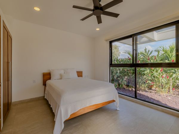 A minimalist bedroom with a large window, a ceiling fan, a bed with white linens, and a view of lush greenery outside the window.