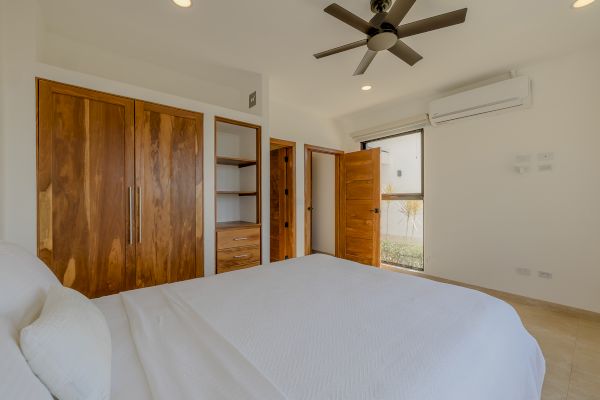 A minimalist bedroom features a white bed, wooden wardrobes, an air conditioner, a ceiling fan, and a window with a view.