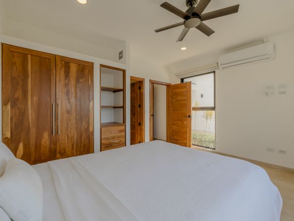 A minimalist bedroom features a white bed, wooden wardrobes, an air conditioner, a ceiling fan, and a window with a view.