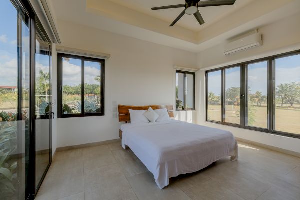 A minimalistic bedroom with large windows, a double bed, white linens, a ceiling fan, and ample natural light entering the room.