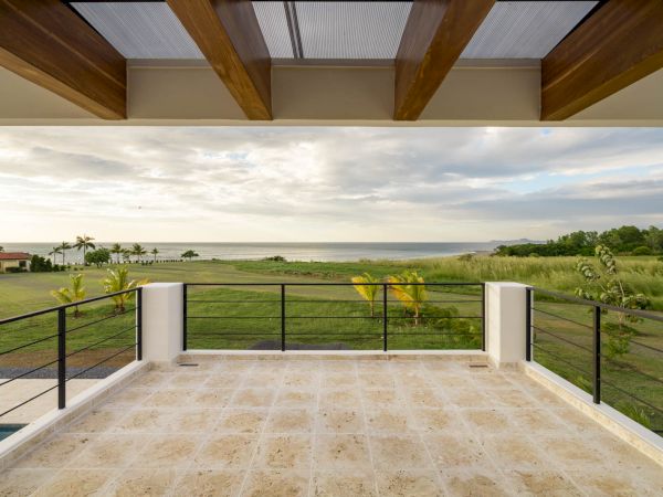 A spacious terrace overlooking a grassy field with palm trees, and a view of the ocean under a partly cloudy sky, framed with wooden beams.