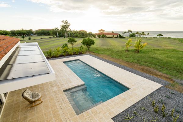 An outdoor pool with a patio area is shown, overlooking a green lawn with trees and a distant view of the sea under a partly cloudy sky ending the sentence.