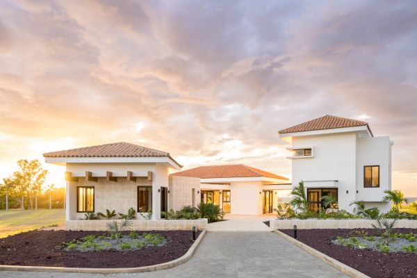 A large, modern house with a courtyard entrance, surrounded by a landscaped garden and set against a dramatic sky with a sunset glow ending the sentence.