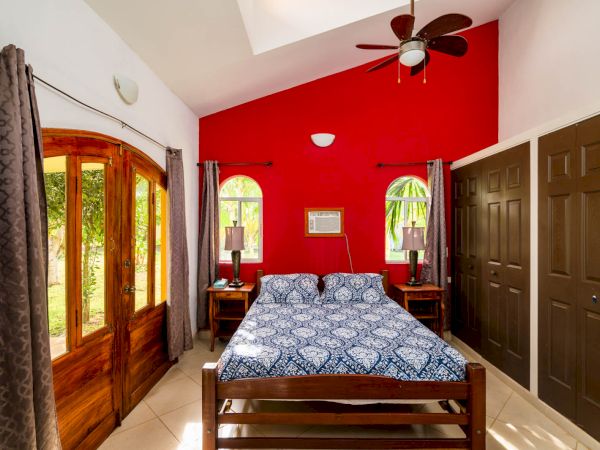 The image shows a bedroom with a red accent wall, a bed in the center, two nightstands, a ceiling fan, large wooden doors, and closets.