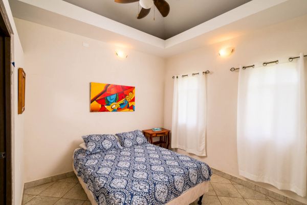 A bedroom with a bed, blue patterned bedding, colorful painting, two windows with white curtains, a small table, and a ceiling fan.