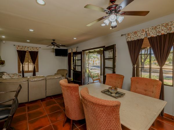 A cozy living room and dining area with a sofa, dining table, ceiling fans, and large windows with curtains, featuring a warm and inviting atmosphere.