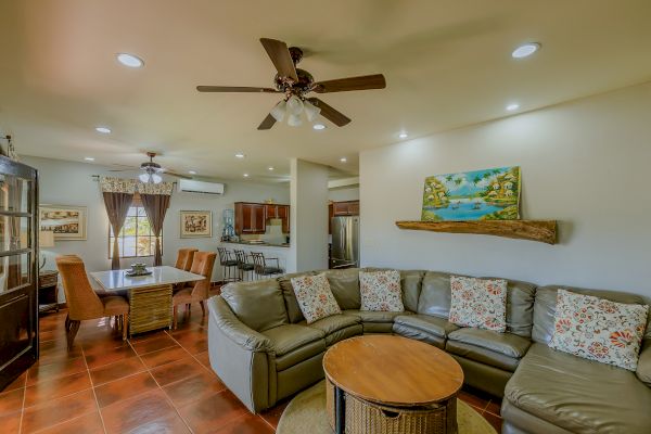 A cozy living room with a sectional sofa, colorful cushions, a round coffee table, ceiling fan, dining area, and kitchen in the background.