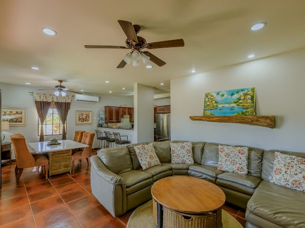 A cozy living room with a sectional sofa, colorful cushions, a round coffee table, ceiling fan, dining area, and kitchen in the background.
