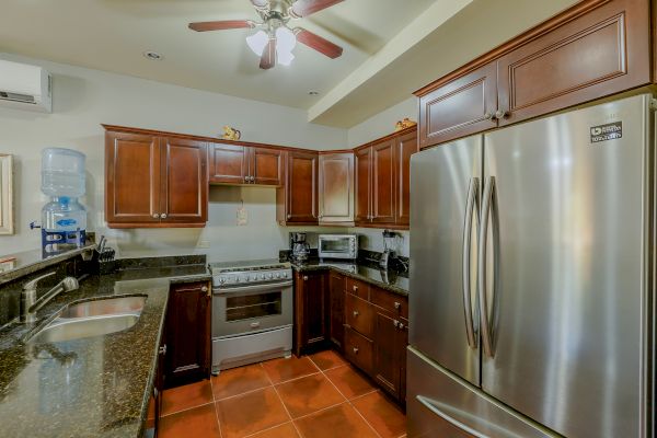 This image shows a modern kitchen with wooden cabinets, stainless steel appliances, and a ceiling fan. There is a water dispenser on the countertop.