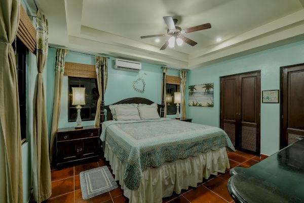 A cozy bedroom with a ceiling fan, a bed with blue bedding, two bedside lamps, curtains, and a beach-themed painting on the wall.