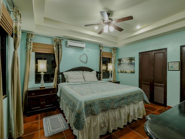 A cozy bedroom with a ceiling fan, a bed with blue bedding, two bedside lamps, curtains, and a beach-themed painting on the wall.