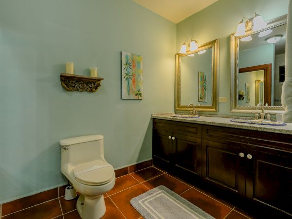 The image shows a bathroom with a toilet, double sink vanity, large mirror, wall décor with candles, artwork, and two wall-mounted lights.