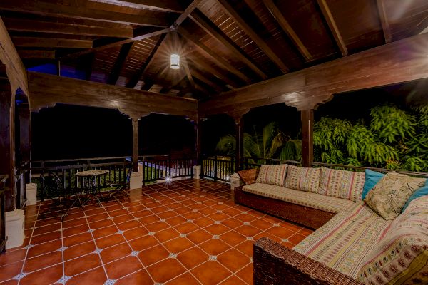 An outdoor patio with a wooden ceiling, hanging light, tiled floor, wicker sofa with cushions, table, chairs, and lush greenery in the background.