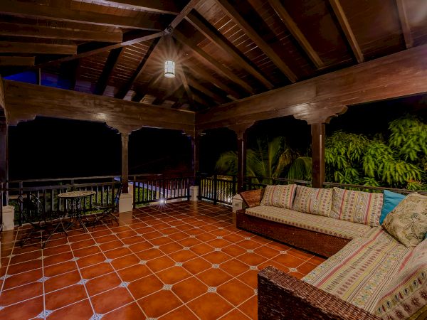 An outdoor patio with a wooden ceiling, hanging light, tiled floor, wicker sofa with cushions, table, chairs, and lush greenery in the background.