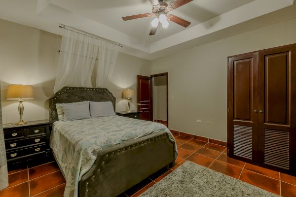 A bedroom with a bed, patterned bedspread, two bedside tables with lamps, a ceiling fan, and a wooden wardrobe on a tiled floor, ending the sentence.