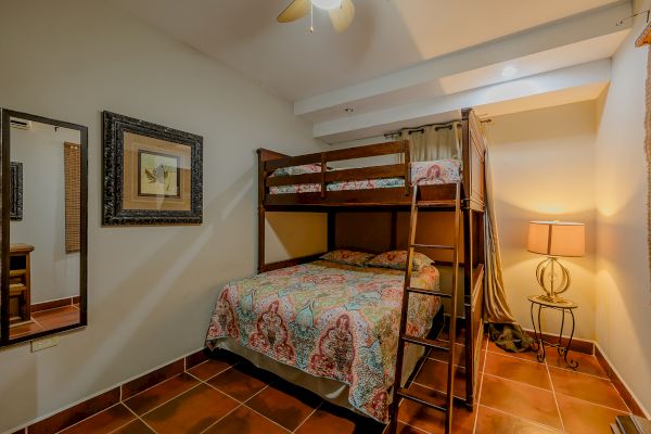 This image shows a small bedroom with bunk beds, a framed picture, a floor lamp, a large mirror, and tiled flooring, all warmly lit.