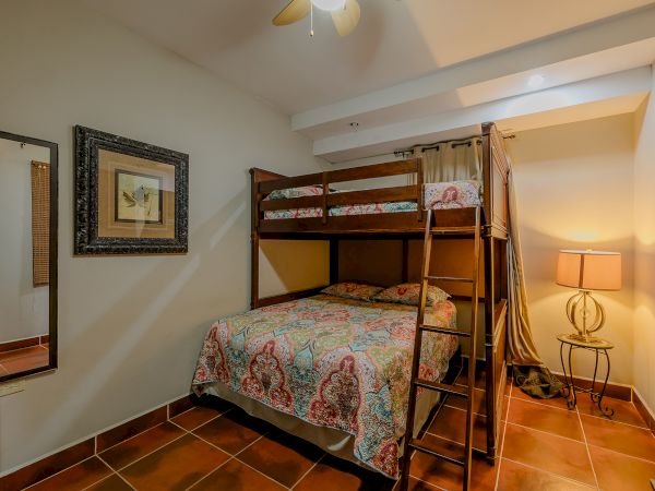 This image shows a small bedroom with bunk beds, a framed picture, a floor lamp, a large mirror, and tiled flooring, all warmly lit.